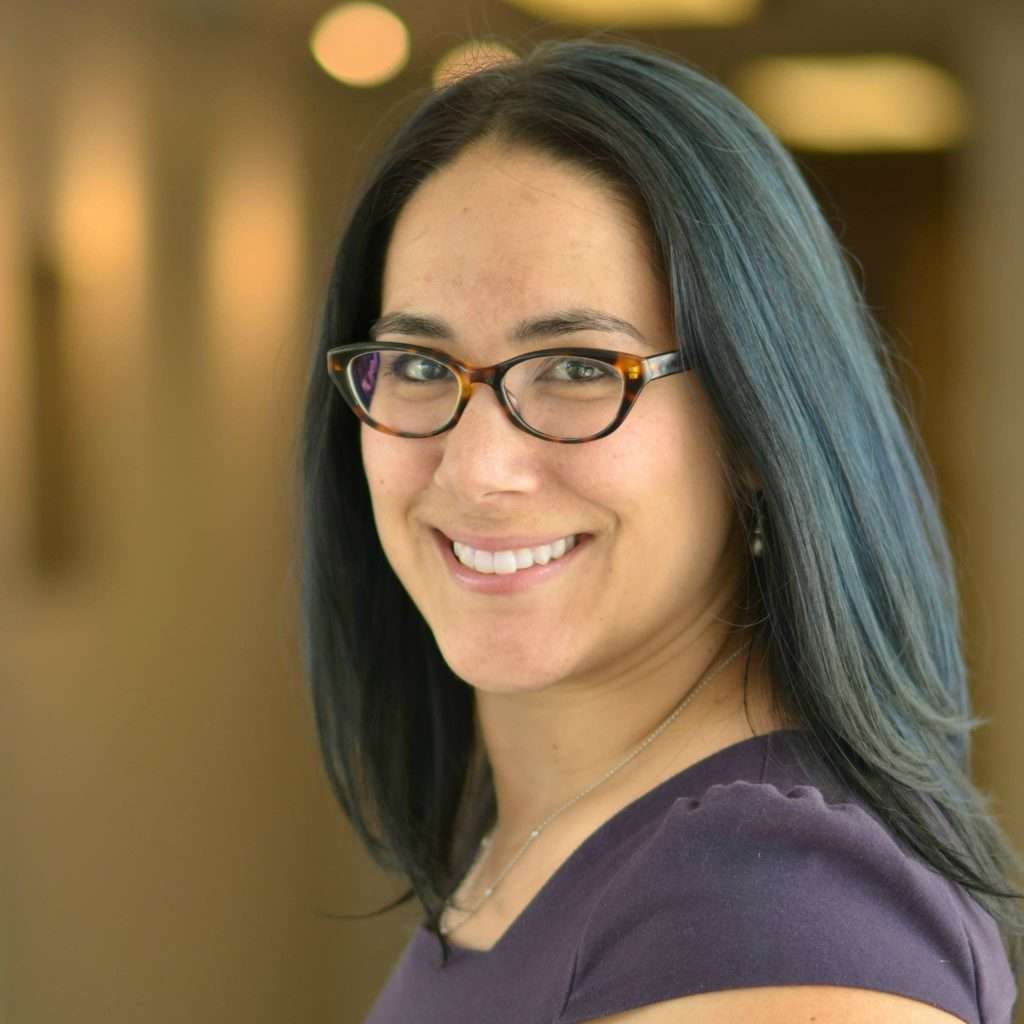 Fond flou. Femme aux cheveux noirs, portant tes lunettes, qui regarde vers la caméra en souriant