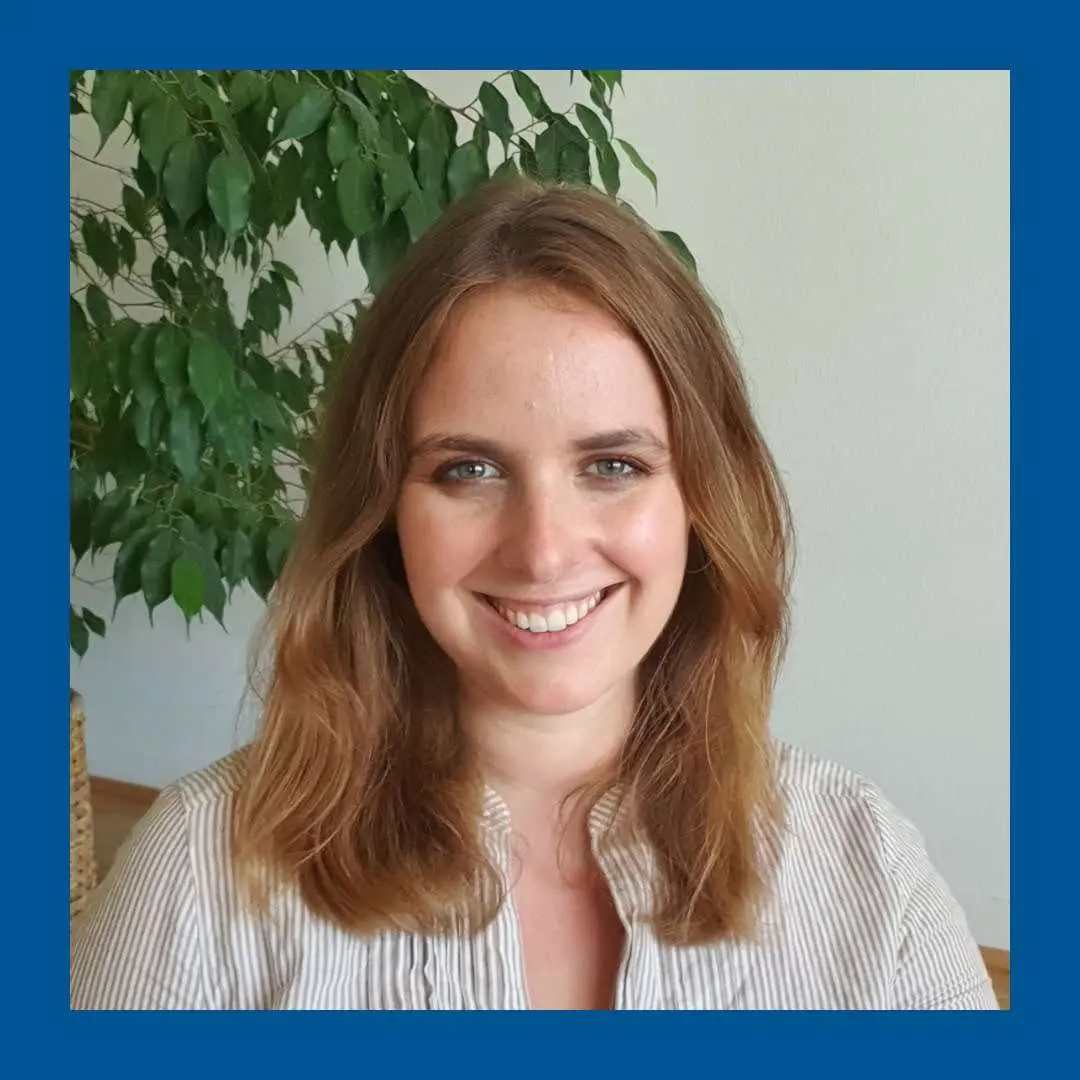 Blue frame. Blonde woman in front of grey wall and ficus smiling towards camera.