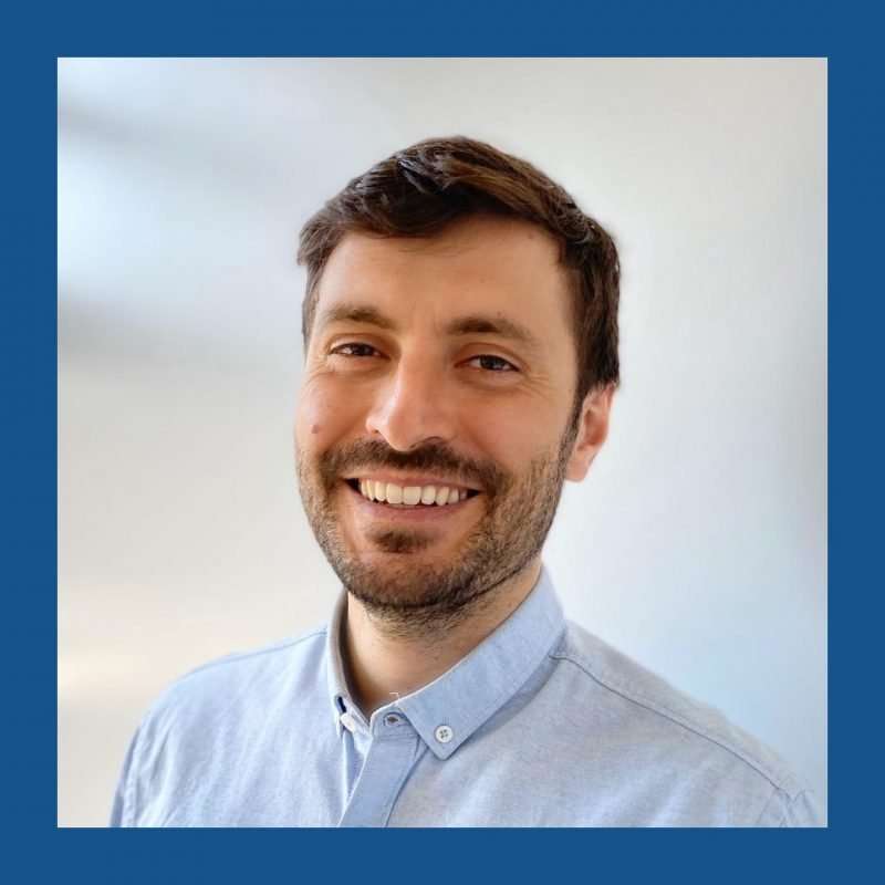 Dark haired man with short beard smiling at camera. Off-white backrgound and blue frame.