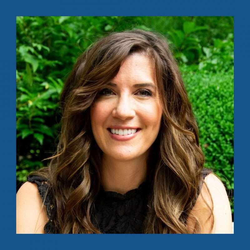 Woman with long brown hair wearing black sleeveless shirt looking and smiling towards camera, with a backdrop of green shrubs