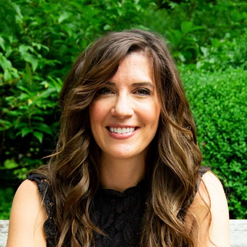 Woman with long brown hair wearing black sleeveless shirt looking and smiling towards camera, with a backdrop of green shrubs