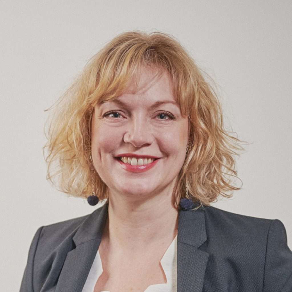 Femme blonde aux yeux bleus regardant et souriant vers la caméra.