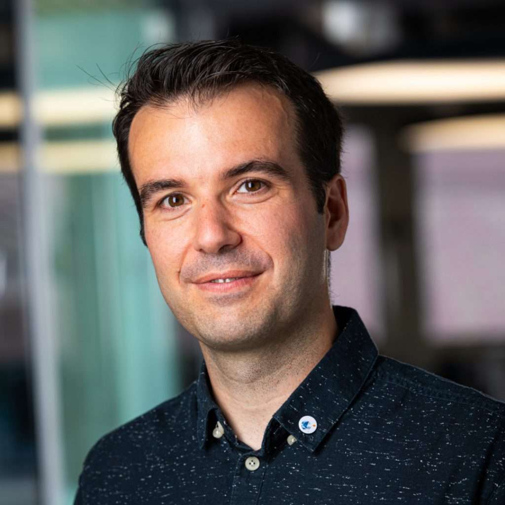 Dark-haired man in blue shirt looking and smiling at the camera. Blurry background