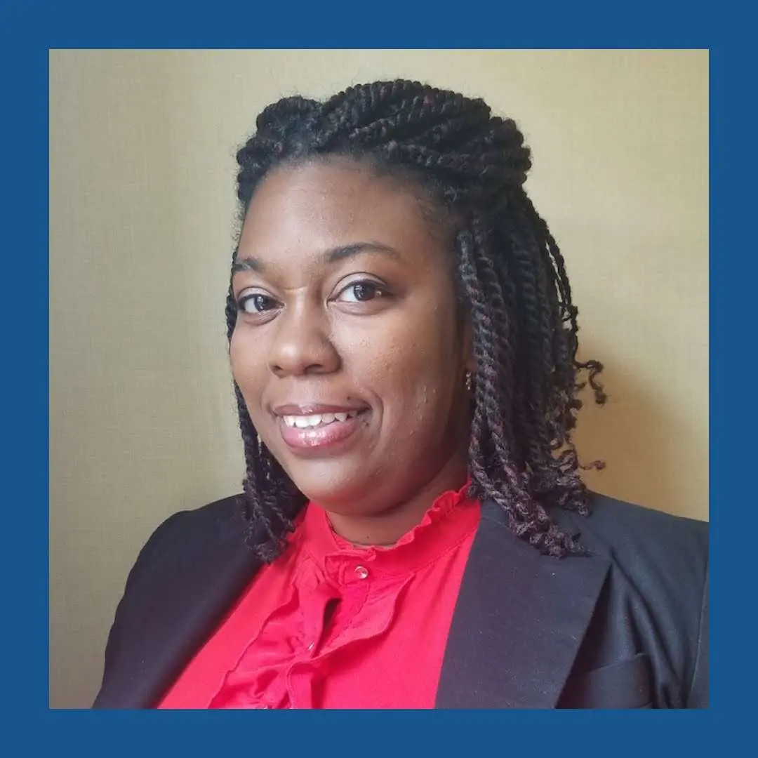 Black woman with braided hair, wearing a red shirt and dark blue blazer, looking and smiling at the camera. Navy blue frame.